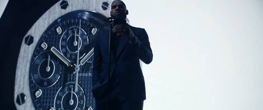 Person in suit speaks on stage with a large watch image behind.