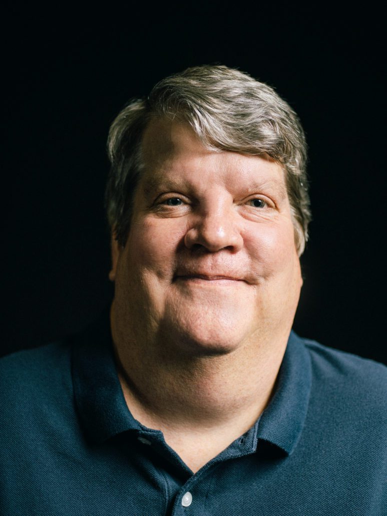 Smiling man in a dark shirt against a black background.