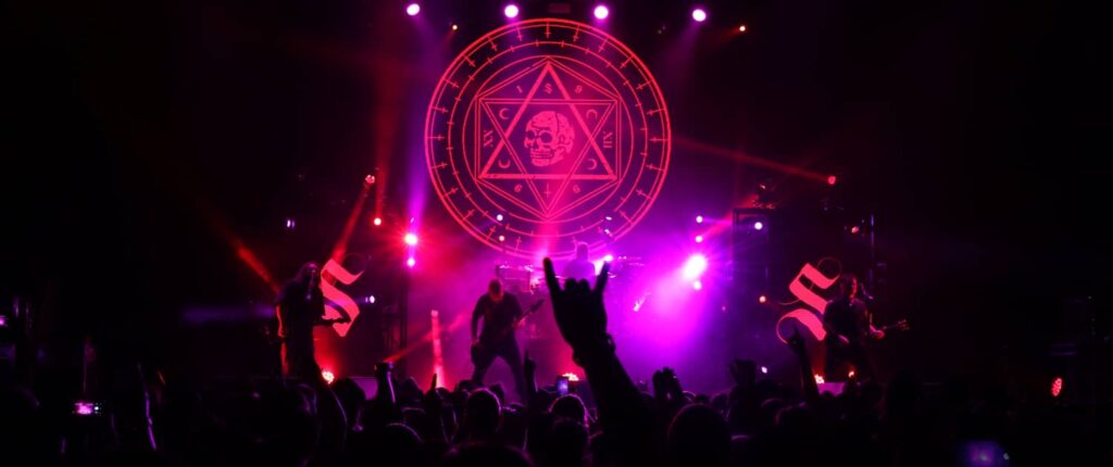 Rock band performing on stage with purple lighting and geometric backdrop.