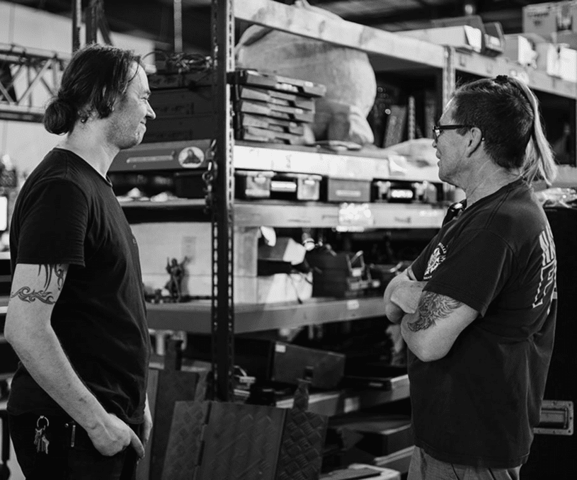 Two men with tattoos talking in a warehouse setting.